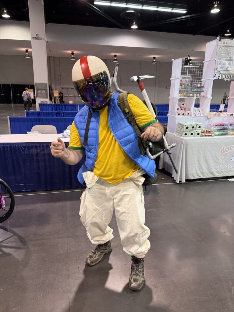 cosplayer in a yellow shirt, blue puffer vest, white cargo pants, and a red-and-white helmet with mirrored visor, carrying a pickaxe prop on the convention floor in Anaheim