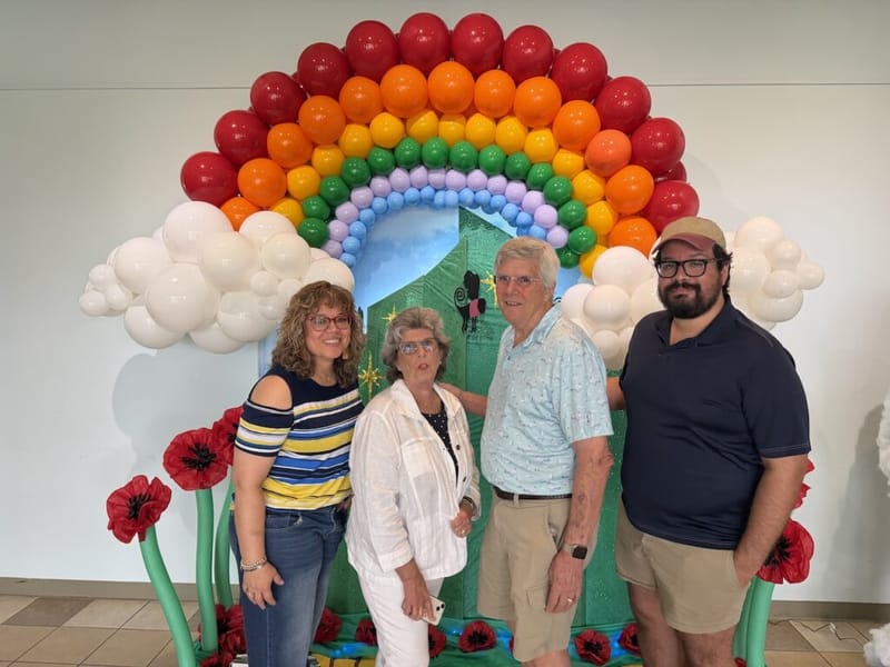 Four people posing in front of a large rainbow balloon arch with decorative red poppy flowers at the base