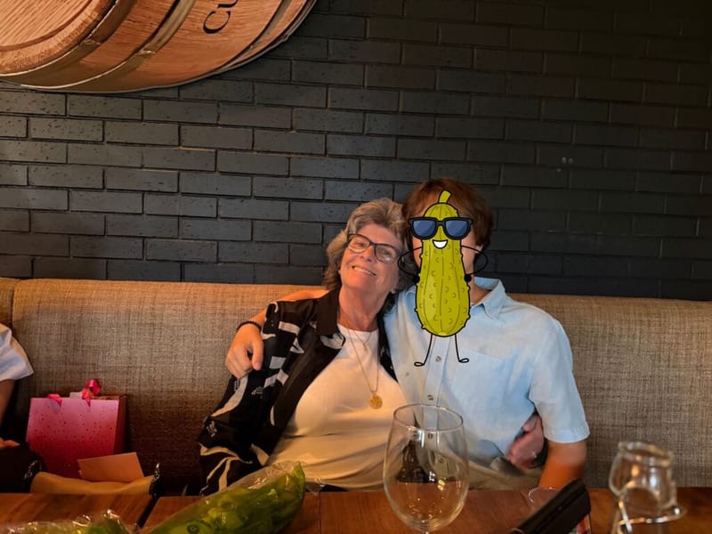Two people seated at a restaurant booth, one with a cartoon pickle sticker covering their face, posing and smiling