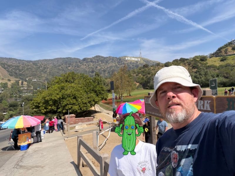 John W. Brandes taking a selfie at Lake Hollywood Park with the Hollywood Sign visible on the hills behind him, a cartoon pickle sticker overlaid in the foreground